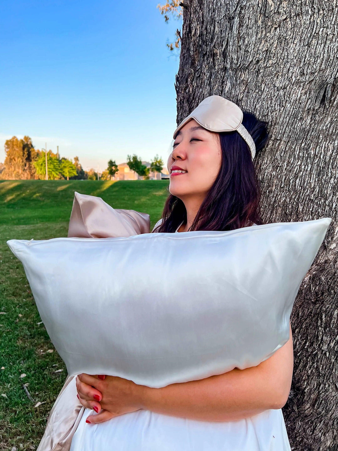 Woman in white showcasing a champagne silk sleep mask outdoors near a tree